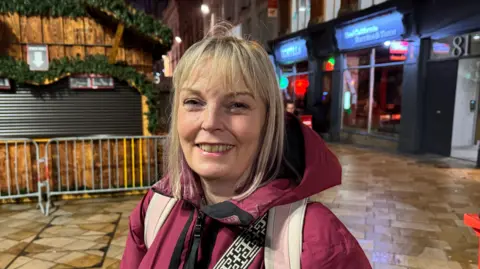 A woman in a red puffer coat is standing in Birmingham city centre near the Christmas market and smiling at the camera
