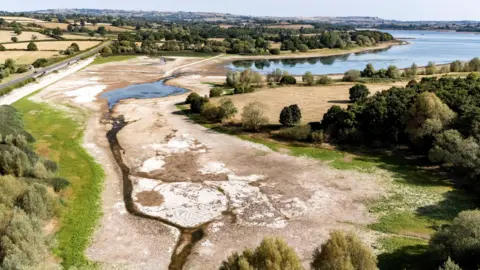 PA Media A drone shot of Chew Valley Lake showing the water line receding around 100 metres from its usual line and exposing dry mud. It is a bright sunny day and the grass in the fields is yellowed