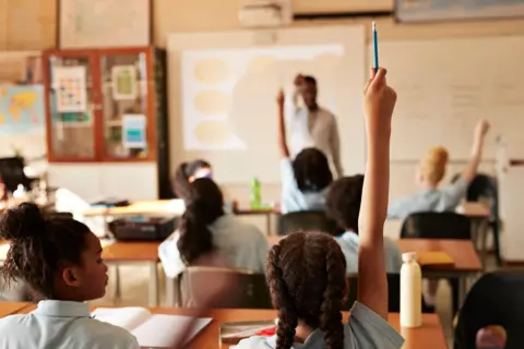 Klaus Vedfelt/Getty Students seated at desks in a classroom, with several raising their hands holding pens. Open notebooks and water bottles are visible on desks. At the front, a teacher stands near a whiteboard displaying a projected presentation with circular diagrams.
