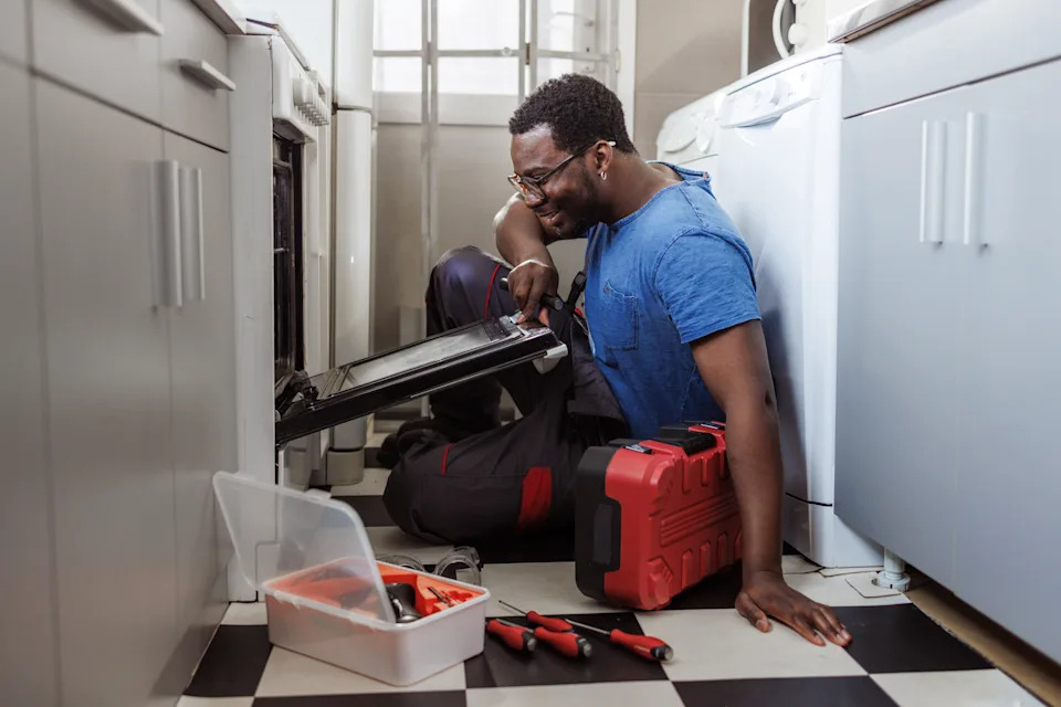 A person in work attire repairs an oven in a kitchen, surrounded by tools, illustrating a home appliance service scenario