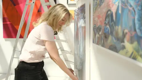 Irina Mackie Ruth Millington stands with her hands under a painting displayed on a gallery wall. There are stepladders behind her as the exhibition is put together.
