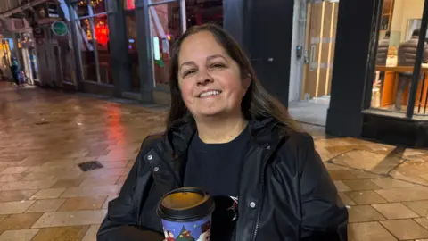 A woman in a black coat and black T-shirt is holding a disposable coffee cup and smiling at the camera in front of some shop store fronts