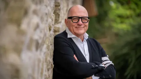 Animo TV 2025 Hugh Wallace standing against a stone wall with his arms folded. He is bald, wearing glasses, a black cardigan with white shirt underneath.