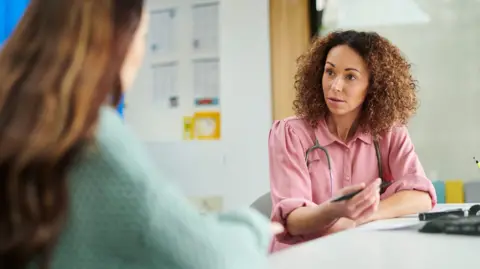 Getty Images A female GP wearing a pink blouse and a stethoscope around her neck, talks to a female patient with long, brown hair (who is blurred and the back of her head is visible only in the foreground)