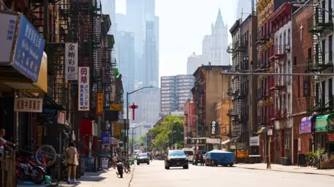 Getty Images Chinatown street in New York City during the day featuring historical buildings with fire escapes and modern skyscrapers in the background.