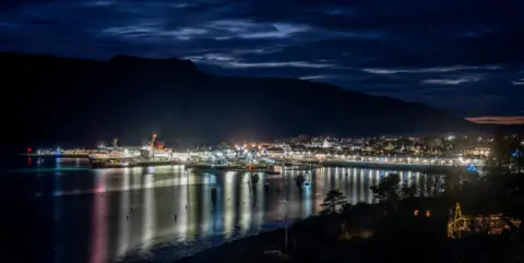Sam Bilner A nigh view of Ullapoool Harbour with lights reflected in the water. A ferry is berthed to the left of the picture. 