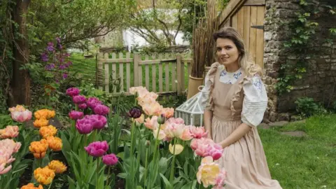 Ramona Jones A woman wearing a brown frilly dress with a white puff sleeve shirt is kneeling in front of tulips in a garden. The tulips are all different colours, orange, purple and pink, and behind her is a lush garden.
