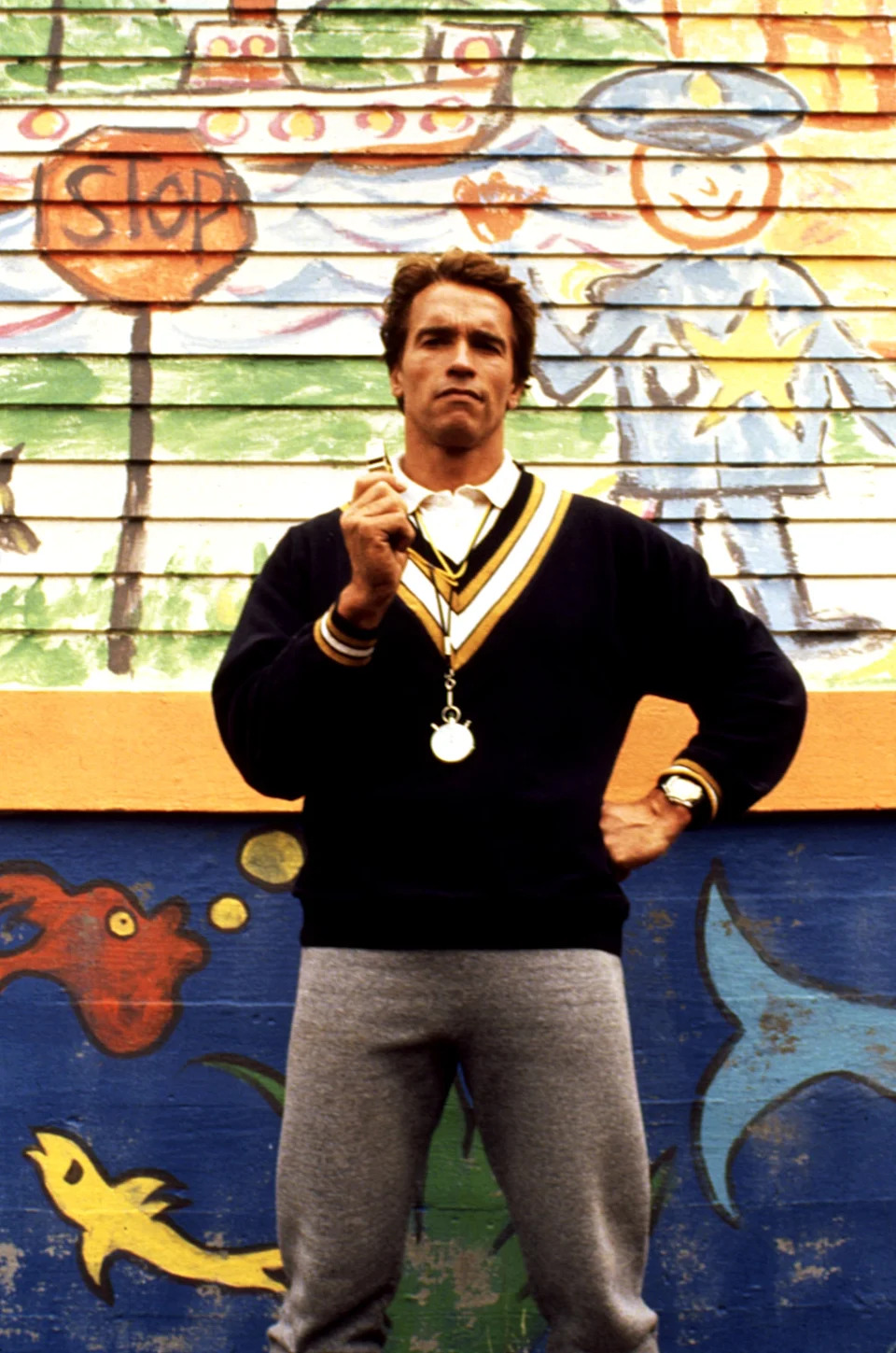 Person standing in front of a colorful mural wearing a casual outfit with a sweater and sweatpants, holding a whistle, exuding a sporty vibe