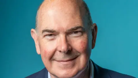 GMICB A bald, older man wearing a navy blue suit smiles against a lighter blue background.