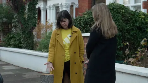 Alison Murphy is wearing a yellow coat and gestures at the stump of the tree while speaking to a reporter in a black coat. A white wall is in the background.