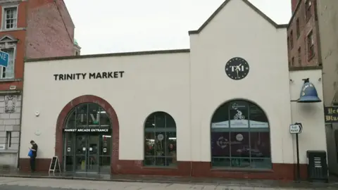 An early 20th century market hall building  with an arched entrance and two matching windows. It is painted cream with red brick details. A round black clock face doubles as a sign with the letters "T M" printed on it. Another sign reads "Trinity Market". To the right, a large blue bell advertises the Blue Bell pub.