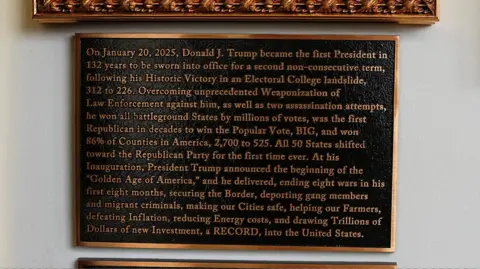 Reuters A close up of the text on Trump's plaque. It includes the phrase "he delivered, ending eight wars in his first eight, securing the Border, deporting gang members and migrant criminals, making our Cities safe, helping our Farmers, defeating inflation..."