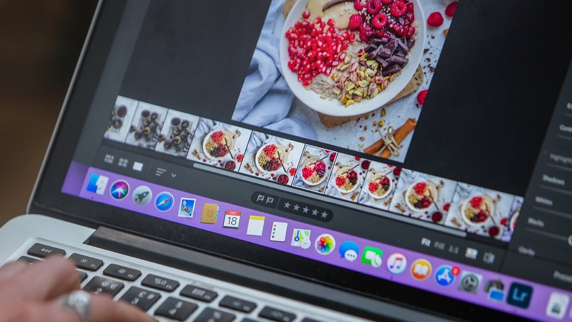 A laptop screen displays a close-up of a bowl of oats topped with red raspberries, pomegranate seeds, chocolate, and pistachios within an Adobe Lightroom interface