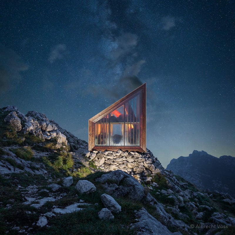 A small, modern glass cabin with a sharp angled roof sits on rocky terrain under a starry night sky, surrounded by mountains. The cabin's warm interior glows through its transparent walls.