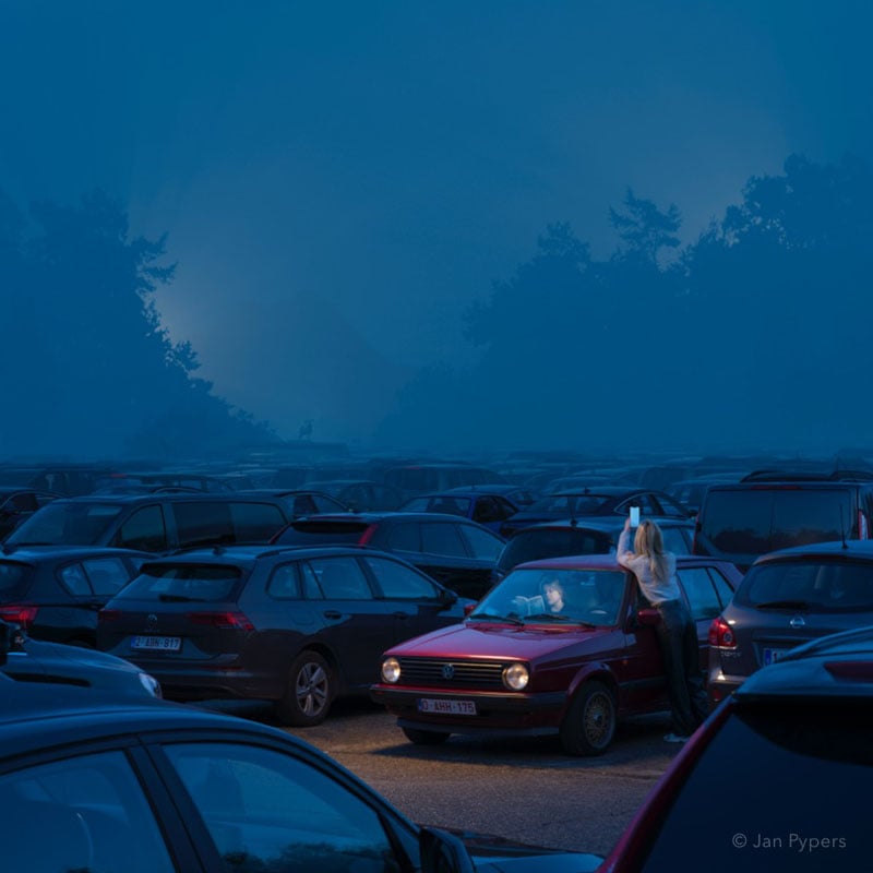 A woman stands outside a red car in a dimly lit parking lot at dusk, using her phone. The car’s headlights are on, and the lot is filled with other parked vehicles under a misty blue sky.