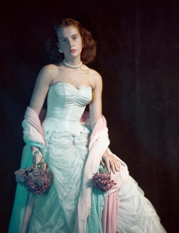 A young woman in a strapless white gown with a pink shawl draped over her arms holds bouquets of pink flowers. She wears a pearl necklace and gazes softly forward, posed against a dark background.