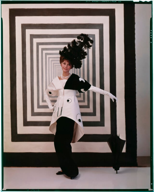 A woman poses in front of a black and white geometric square backdrop, wearing an elaborate feathered hat, pearl necklace, black and white dress with circular patterns, long white gloves, and holding a closed black umbrella.