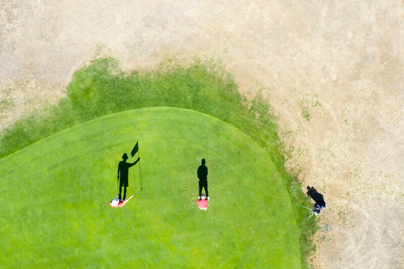 Aerial view of two people on a golf green with bright shadows and golf bags nearby. The lush green contrasts with the dry, brown grass surrounding the area.