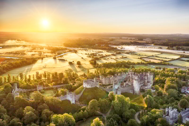 Aerial view of a historic castle surrounded by lush green trees and fields at sunrise, with golden sunlight and morning mist illuminating the rural landscape.