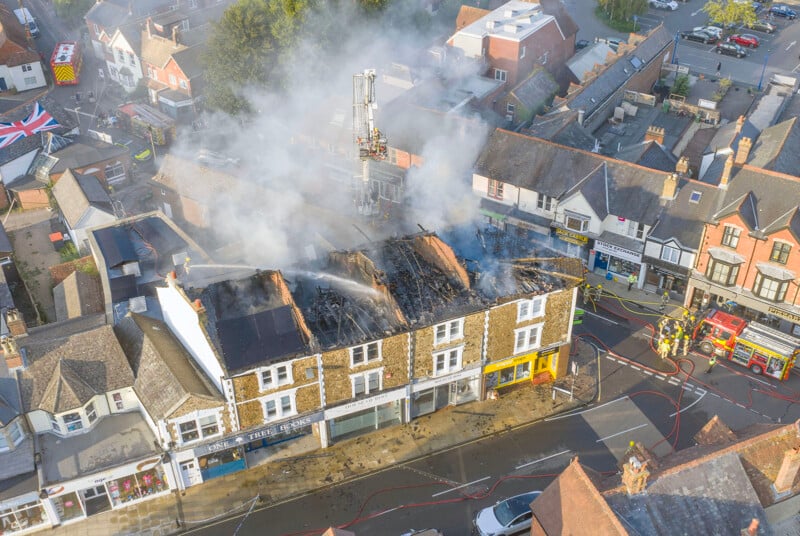 Aerial view of a fire-damaged building with firefighters and fire trucks on the street below. Thick smoke rises from the partially destroyed roof as emergency crews work to control the scene. Nearby buildings and parked cars are visible.