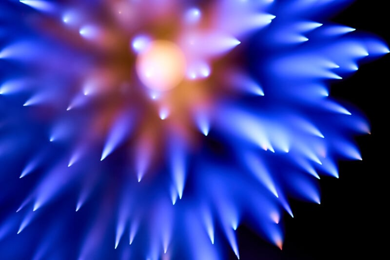 Abstract image featuring a burst of sharp, blue and purple spikes radiating from a bright central point, set against a dark background. The overall effect resembles a glowing, spiky flower or a starburst pattern.