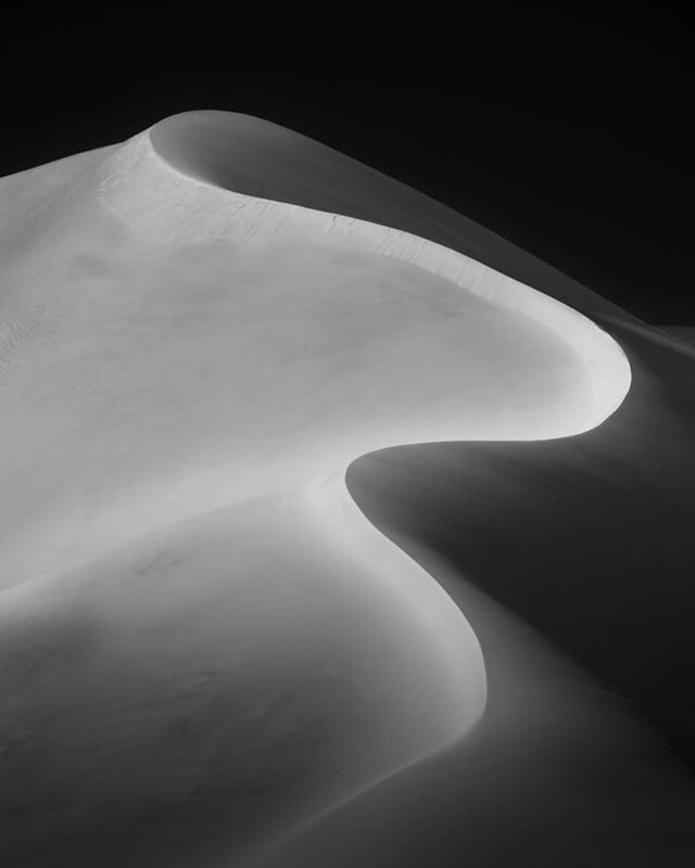 Black and white photo of smooth sand dunes with soft, curving lines and dramatic shadows, creating an abstract and minimalist landscape against a dark sky.
