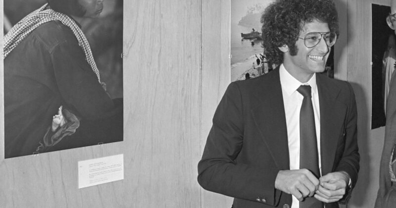 A man in a suit and tie with curly hair and glasses smiles while standing indoors next to a wall displaying black-and-white photographs.