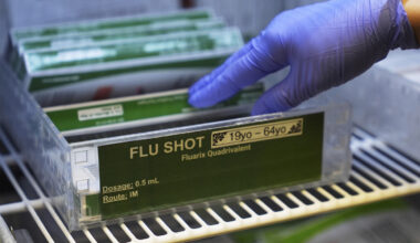 A hand wearing a latex glove selects a flu shot inside a refrigerator.