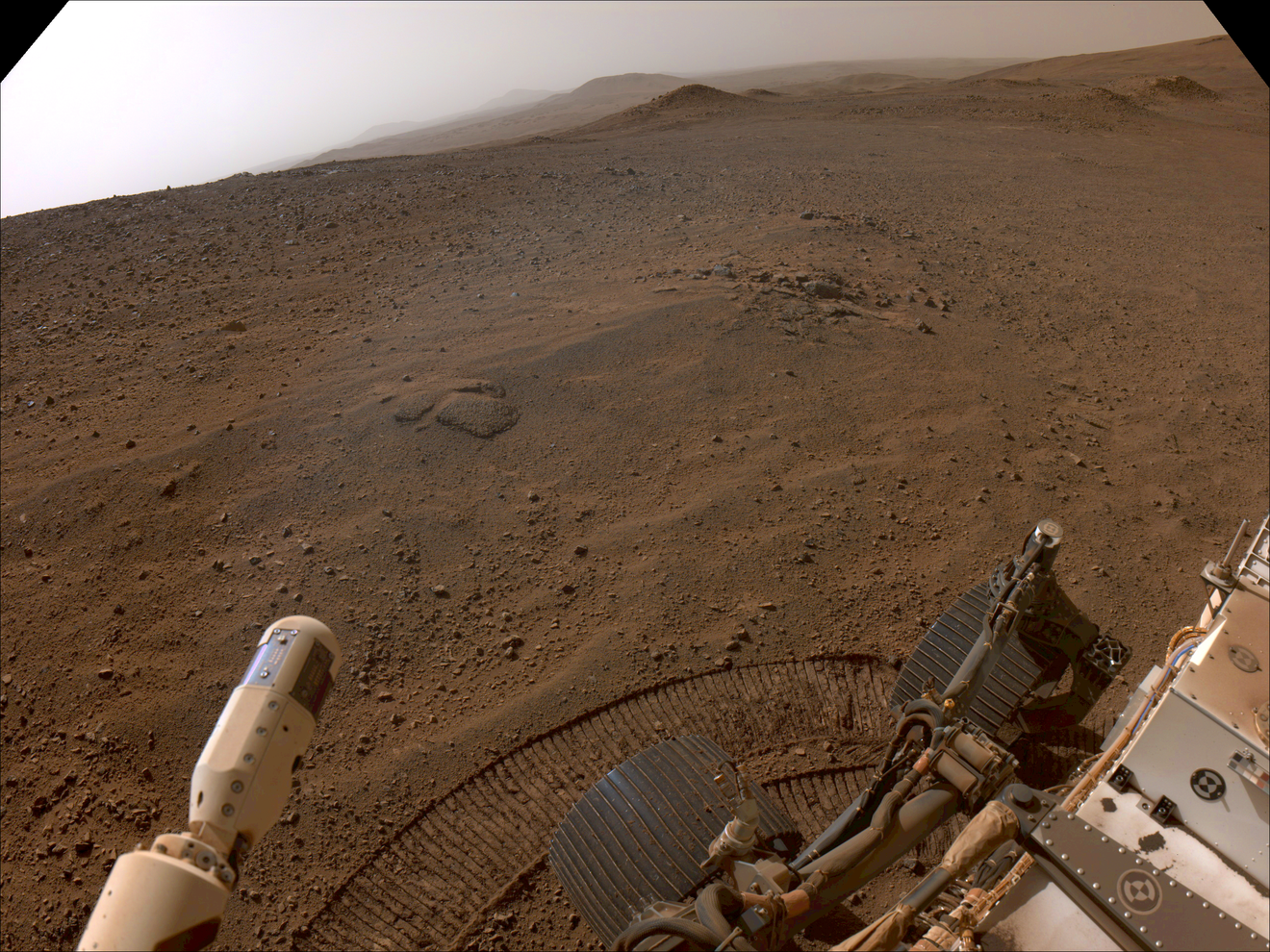 An image taken by Perseverance with two of its wheels in the bottom right corner as it overlooks a cloudy Martian landscape