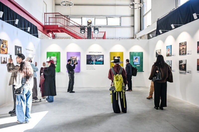 People view and discuss colorful photographs displayed on white gallery walls in a bright, modern exhibition space with high ceilings and red stairs in the background.