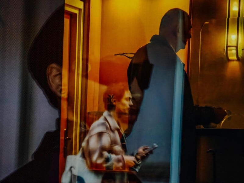 A layered, abstract photo showing silhouettes and reflections of people in warm, indoor lighting. One person appears to be walking while using a phone, while another stands at a counter, both partially overlapping.