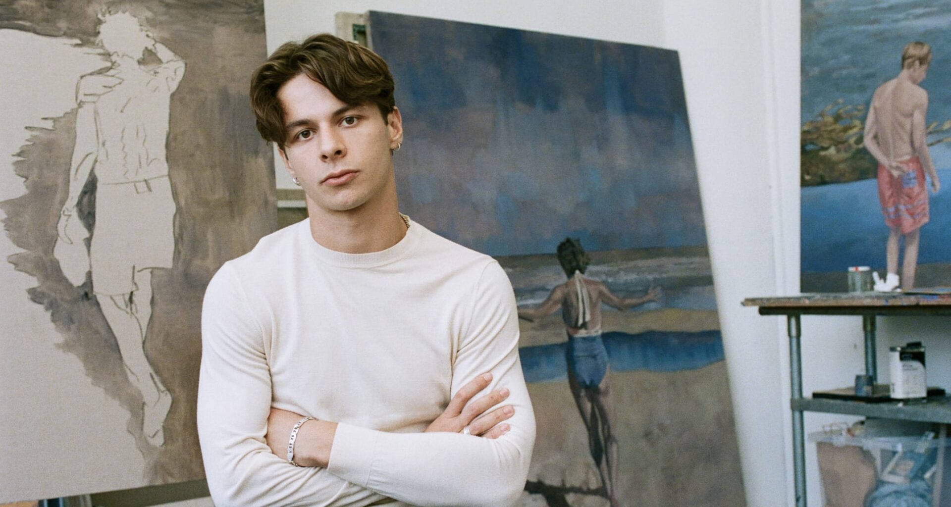 Young man sitting in an art studio with folded arms, surrounded by coastal-themed paintings in various stages of completion.