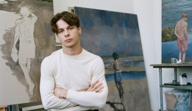 Young man sitting in an art studio with folded arms, surrounded by coastal-themed paintings in various stages of completion.