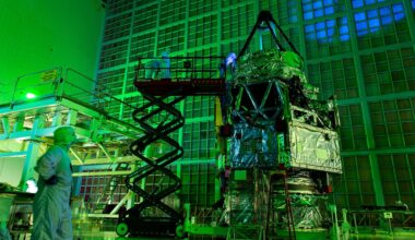 A telescope with a triangular top wrapped in foil stands next to a scaffold with people wearing white clean suits examining it under green light.