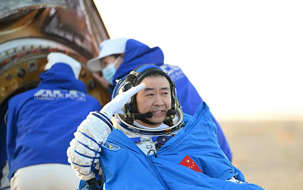 A man wearing a white spacesuit and open helmet with a blue cape on over his gear with a red flag with yellow stars salutes someone off camera to the right of the image.