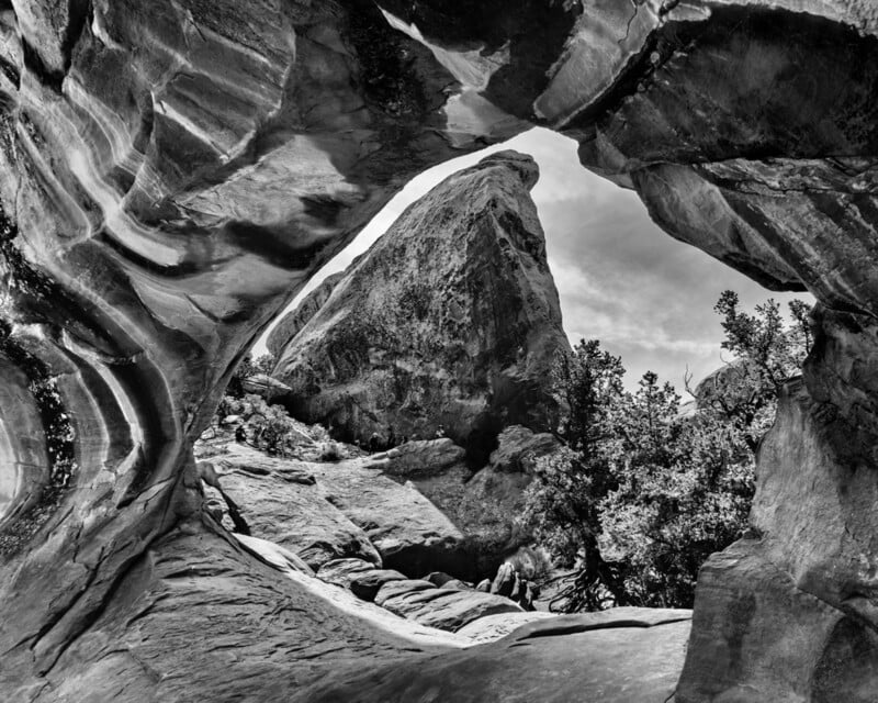 Black and white photo of a rocky arch framing a jagged mountain peak and trees under a partly cloudy sky, capturing a dramatic natural landscape.
