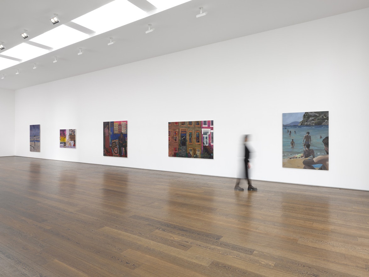A person walking through a spacious art gallery with several colorful paintings displayed on white walls and a wooden floor.