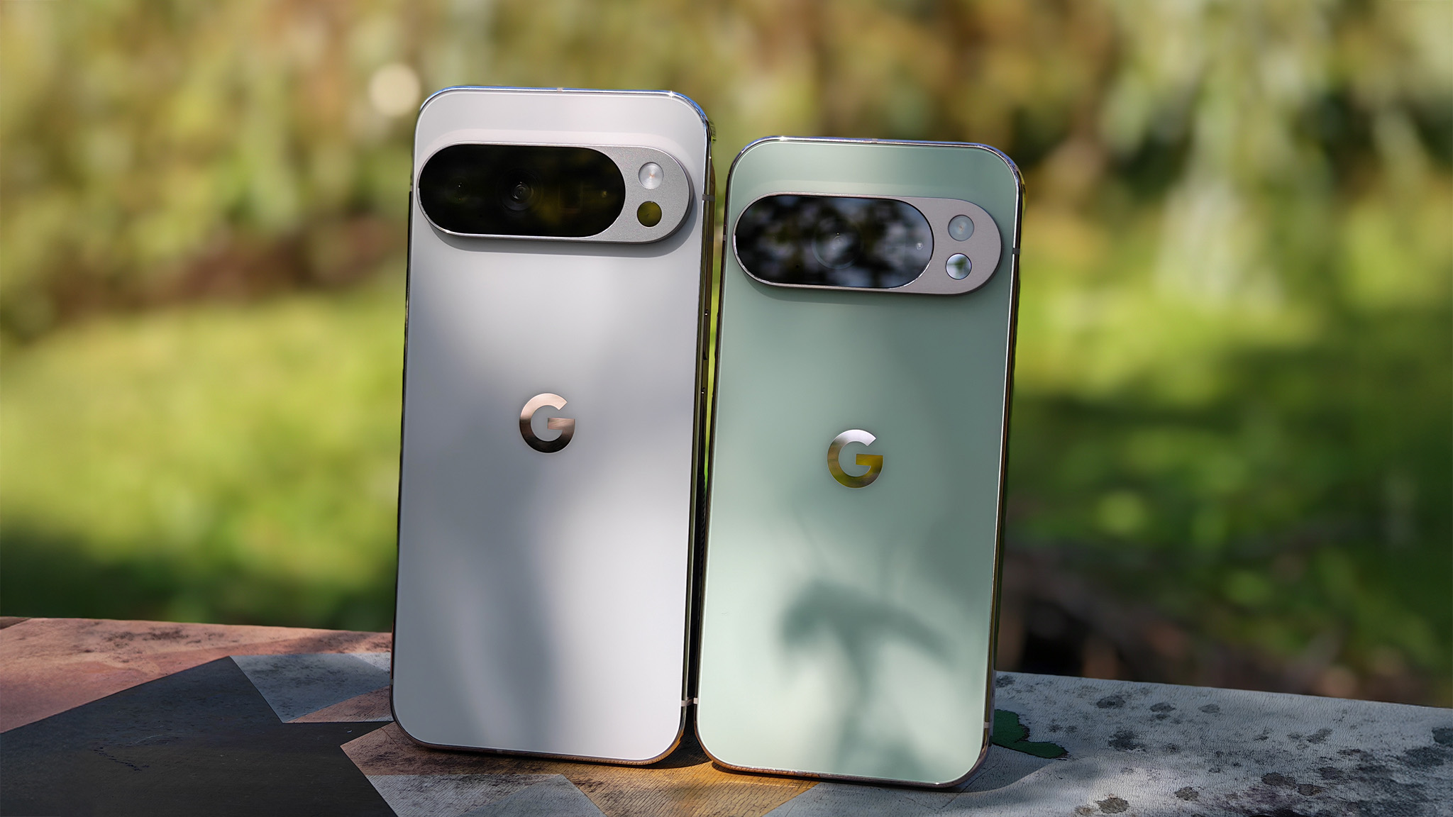 The backs of the porcelain Google Pixel 10 Pro XL and the jade Google Pixel 10 Pro standing next to each other on a bench