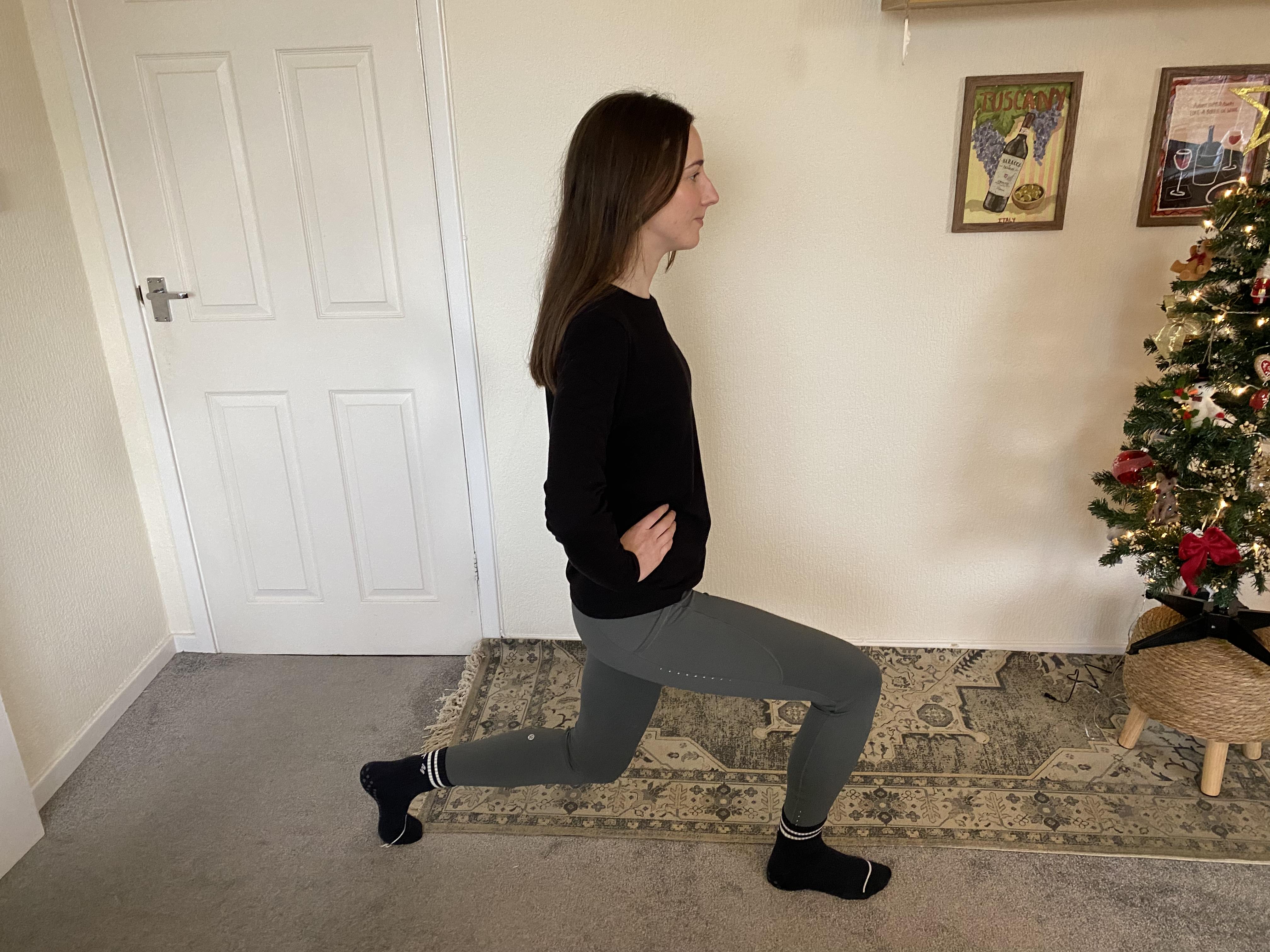 Katie Sims performs a lunge at home. Her right leg is stepped forward, her left leg bent behind her, and her hands are on her hips. We see a Christmas tree and framed pictures behind her.