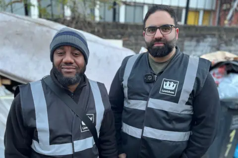 BBC Alain N'Guessan Bi and Eiman Rostami wearing dark Lewisham clothing. They are standing in front of overfilling bins and a stained mattress