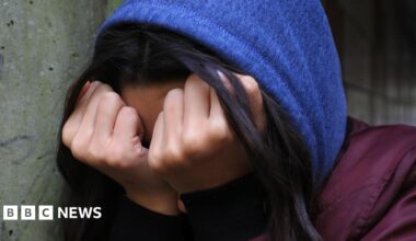 A young woman wearing a blue hooded jumper covers her face with her hands.