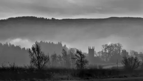 Michael O'Kane A black and white photo of a church in the middle ground, with hills in the background and trees in the foreground.