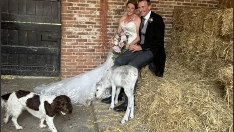 Supplied Angie Nelson is in a white strapless beaded and lace wedding dress, sitting on a hay bale next to her groom, who is wearing a morning suit. The white baby reindeer is leaning against them and a liver and white-coloured springer spaniel is walking across the barn floor.;
