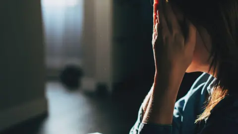 Getty Images A distressed woman sitting on the floor of a dark room with her head in her hands.  She has dark shoulder-length hair and is wearing a blue top.  There is a window in the background.