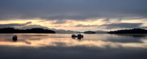 Victor Tregubov Loch Lomond with the sky reflected in the water. Two small fishing boats are in the water and hills are in the background.