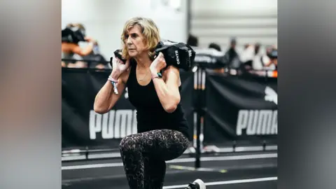 Hyrox Annie Reid during a sandbag lunge walk during a Hyrox race. She rests the sandbag behind her head on her shoulders. She has a concentrated look on her face as she lunges. She wears a black vest top with animal print leggings.