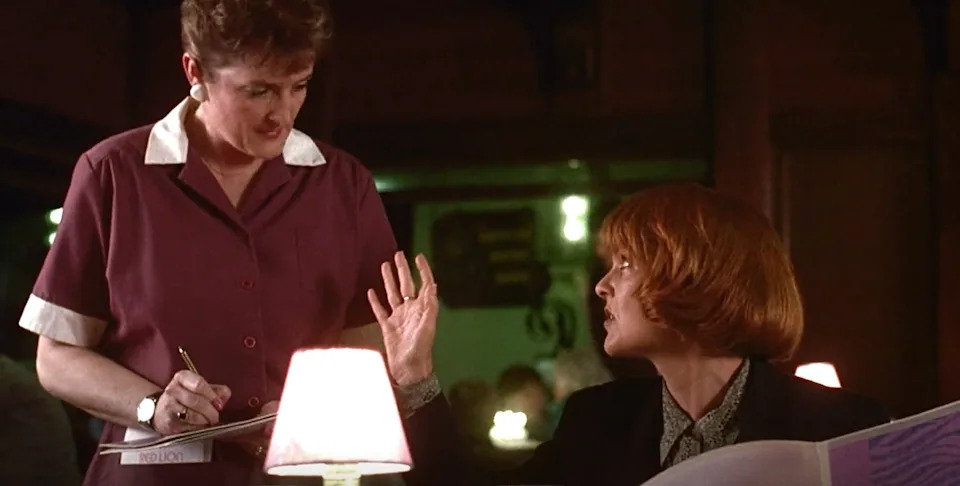 A woman in a dark suit gestures to a waitress with a notepad in a dimly lit restaurant scene