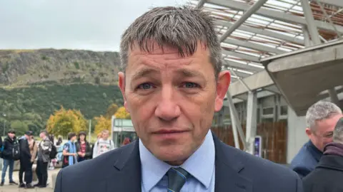 David Threadgold looking straight at the camera. He is wearing a dark suit over a lighter shirt.