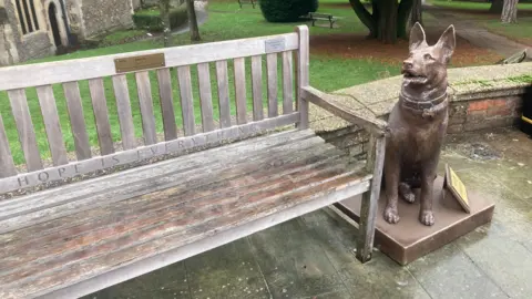 Justin Dealey/BBC A wooden bench with the engraved words “HOPE IS EVERYTHING” on the seat is next to a bronze statue of a German shepherd dog.