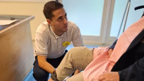NHFT Liam Yeo with a patient, who is sitting down, all you can see if one leg and a body of the patient. Liam is sitting down, wearing a white polo-shirt, has a name badge on and dark trousers, 